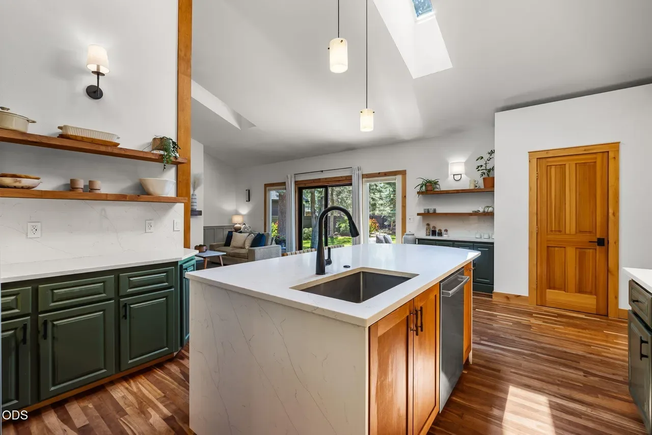 Bright open-plan kitchen with deep green lower cabinets, white quartz island with undermount sink and matte black faucet, pendant lights, vaulted ceiling with skylight and exposed wood beam, and warm wood flooring