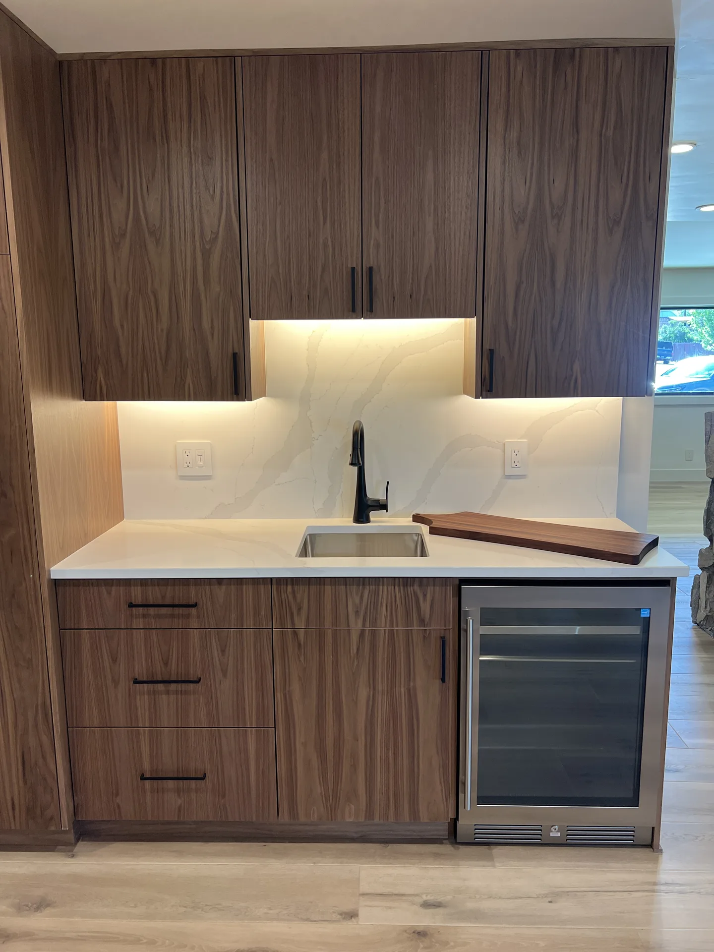 Walnut beverage bar with white quartz countertop and backsplash, undermount bar sink, black faucet and stainless undercounter refrigerator
