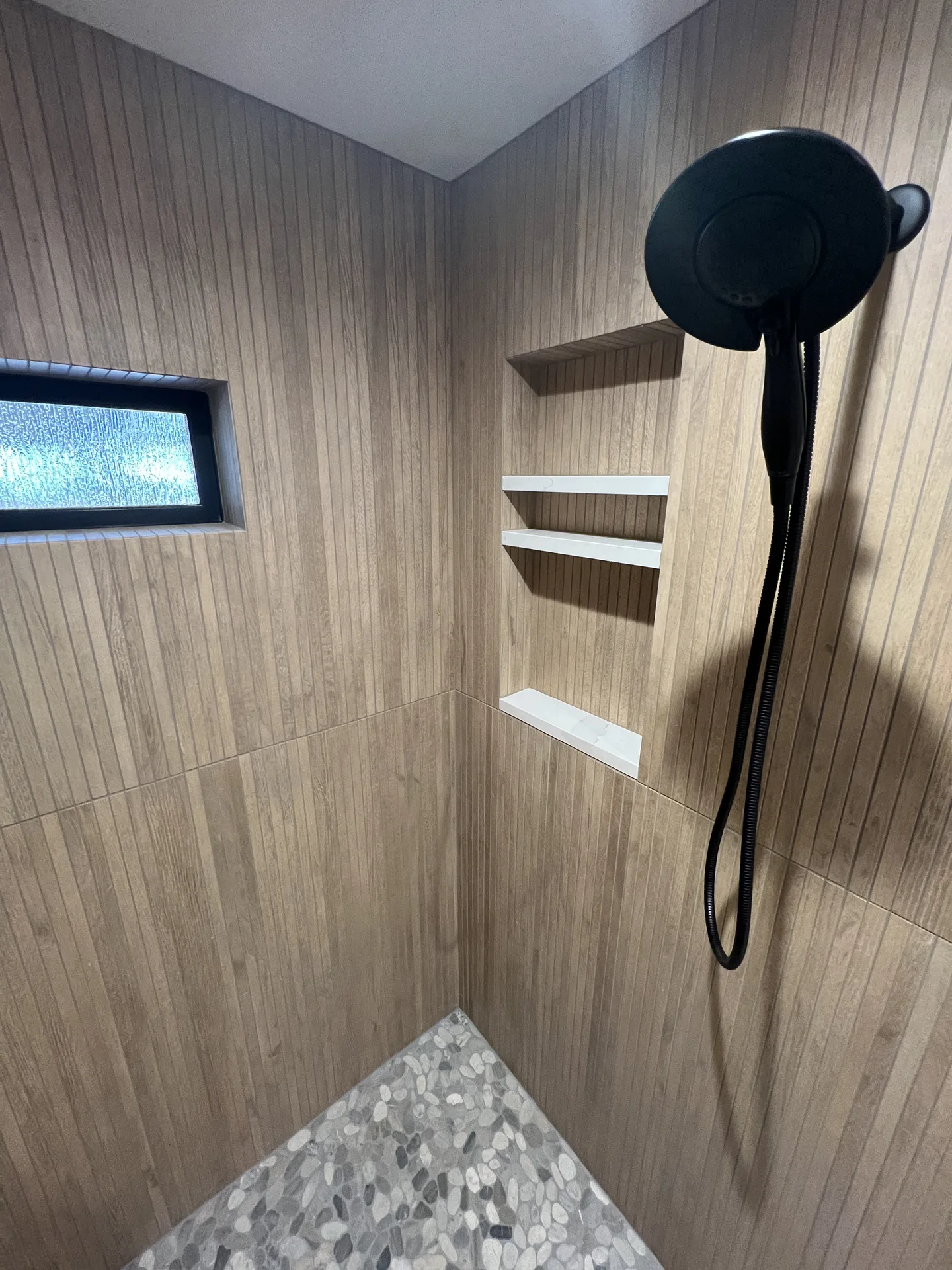 Shower interior with wood-look tile, white quartz niche shelves, black shower head and pebble stone floor