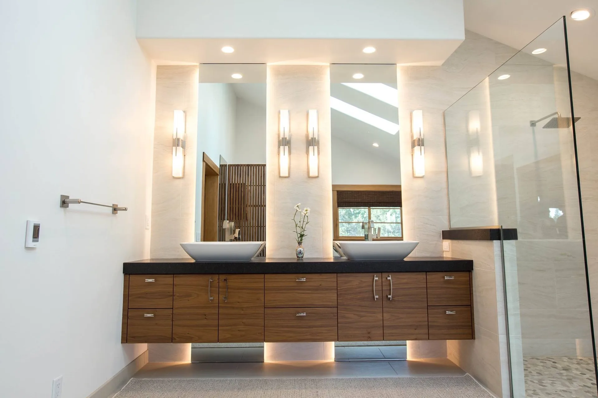 Floating walnut double vanity with dark stone countertop, two white vessel basins, backlit mirrors, vertical sconces, and a glass shower enclosure