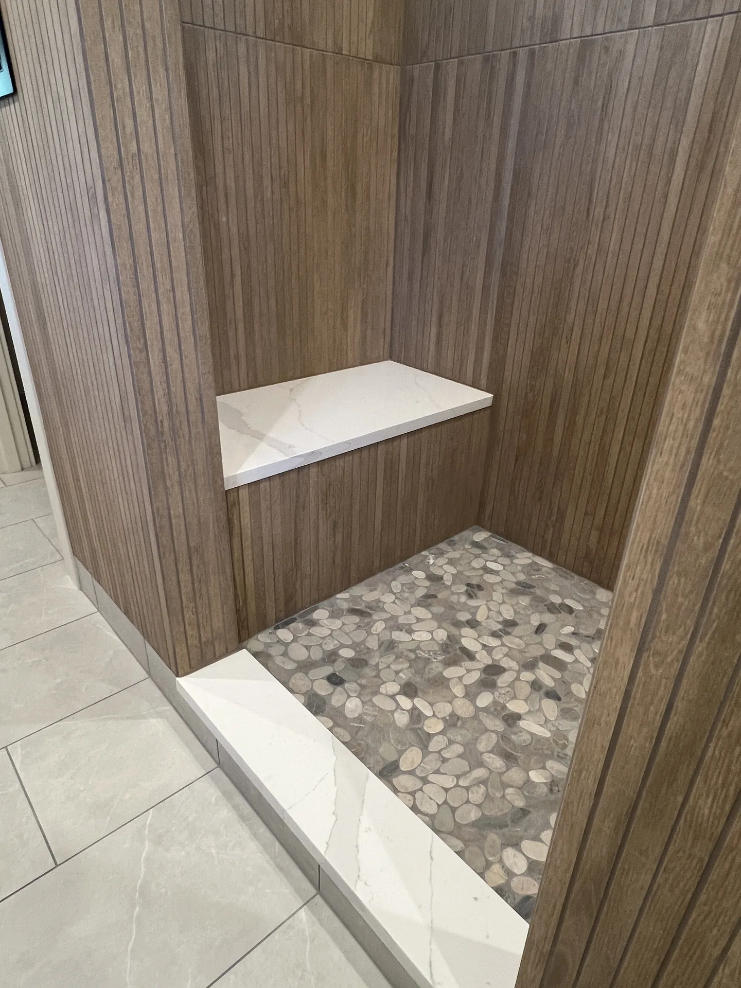 Walk-in shower with wood-look tile walls, a white quartz bench and curb, and a gray pebble stone floor