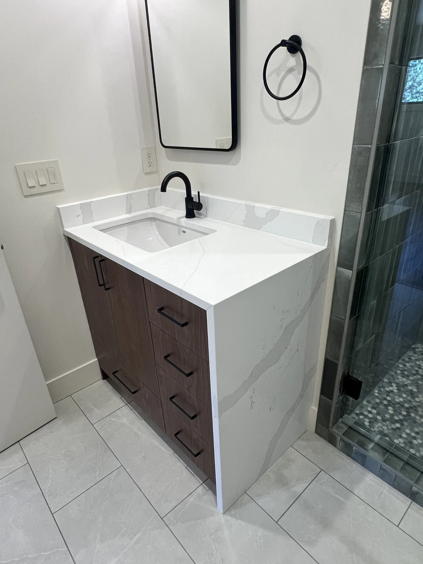 Single bathroom vanity with white quartz countertop and waterfall side panel over dark wood cabinets beside a tiled glass shower
